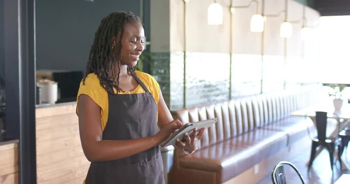 African woman checking tablet after order alert, tapping while facing cafe counter in apron