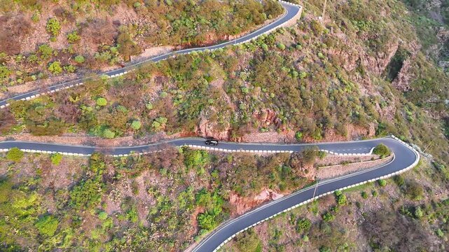 Aerial view of black car driving along a winding mountain road surrounded by lush greenery and rocky terrain, showcasing the scenic landscape and road curvature. Auto trip at scenic route. Journey