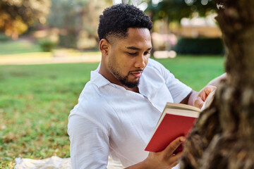 Fototapeta premium Young man student businessman enjoys sunny day while reading a book in park, highlighting his joy and connection to the world around him