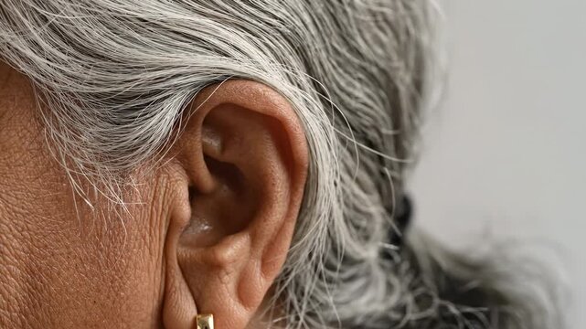 Extreme close up of an elderly woman's ear and gray hair, detailing the wrinkled skin and the earlobe with a golden earring. This shot focuses on aging, hearing problems and audiology concepts