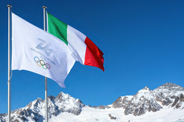 Naklejka premium MILAN, ITALY, DECEMBER 12, 2025. The Italian national flag and the Milano Cortina 2026 Winter Olympics flag with the iconic rings wave over the snowy peaks of the Italian Alps in the sun Ai generated