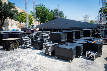 Flight cases and sound equipment on an outdoor cobblestone stage, preparing for a concert event with truss structures and a covered stage setup under a clear sky