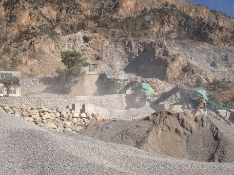 Active open-pit stone quarry in a mountainous region. Industrial landscape featuring deep excavation terraces, rock crushing machinery, and white limestone boulders under a clear blue sky.