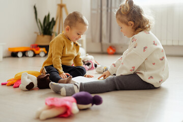 Naklejka premium Two young children sit on the floor with toys around them. They focus on a book laid out in front of them in a well-lit room during the daytime