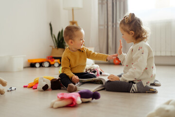 Naklejka premium Two toddlers are sitting on the floor, smiling and playing together. They have toys and a book around them in a bright room with natural light