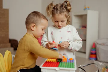 Fotobehang Lego Two children are engaged in building with various colorful blocks inside a well-lit room filled with toys and playful items  © sementsova321
