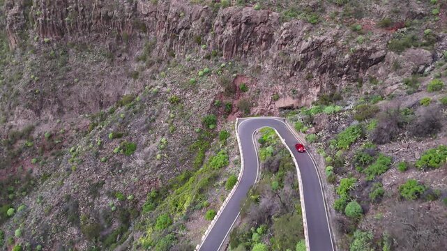 Red car navigates winding mountain road with sharp turns and lush greenery. Auto riding on a scenic route with rocky cliffs and vibrant vegetation. Journey through beautiful nature route. Aerial view
