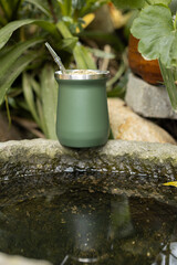 Green stainless steel mate cup with thermos placed outdoors in natural environment. Modern reusable drink container for traditional South American infusion, lifestyle and travel concept.