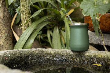 Green stainless steel mate cup with thermos placed outdoors in natural environment. Modern reusable drink container for traditional South American infusion, lifestyle and travel concept.