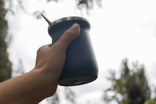 Woman holding green stainless steel mate cup outdoors with blurred natural background. Lifestyle concept of traditional South American drink, wellness, relaxation and sustainable living.