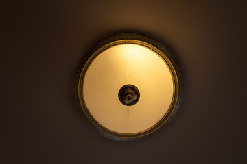 Low angle view of a vintage round ceiling lamp with warm glowing light. Classical interior design element for home or hotel room. Retro style electrical fixture on a dark background.