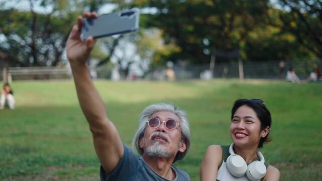 Happy elderly Asian father and daughter smiling on a big grassy esplanade and taking a selfie in the park