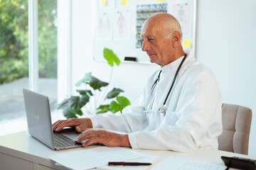 Senior male doctor wearing white uniform with stethoscope working on laptop in his hospital office. Medicine, healthcare and medical