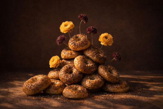 Stack of seeded bagels arranged with small flowers on dark textured surface. Sculptural food composition, warm dramatic light, rustic balance, poetic still life blending nourishment and nature.