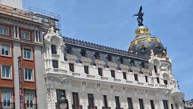 Historic metropolis building with ornate facade and iconic dome topped by a winged statue under a blue sky in Madrid, Spain - 06.13.2025. The image represents urban heritage