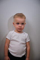 Fototapeta premium Portrait of a Young Boy in a White Shirt Standing Against a Plain Wall, Looking Solemn and Thoughtful