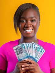 A smiling woman in a pink top holds a fan of blue 100 Ghanaian cedi banknotes against a bright yellow background. The image radiates financial success, joy, and vibrant, high-contrast energy.