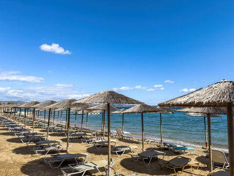 Empty beach loungers under straw umbrellas upon sandy shore within Artemida, Attica, Greece, facing calm Aegean water under bright midday sun and clear blue sky, wide view