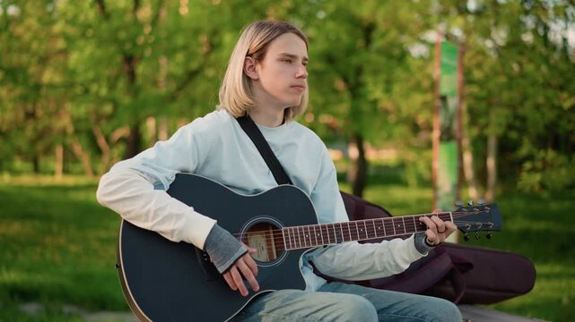 guitarist practicing chords on bench close portrait, focused expression, denim outfit, acoustic black guitar, tuning and strumming, urban park backdrop, candid natural light, indie rehearsal vibe