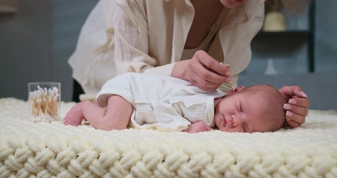 Mother gently cleans sleeping infant's ear with cotton swab on soft blanket, hygiene care. Mother gently cleans newborn baby's ear with cotton swab while baby sleeps peacefully on soft blanket.