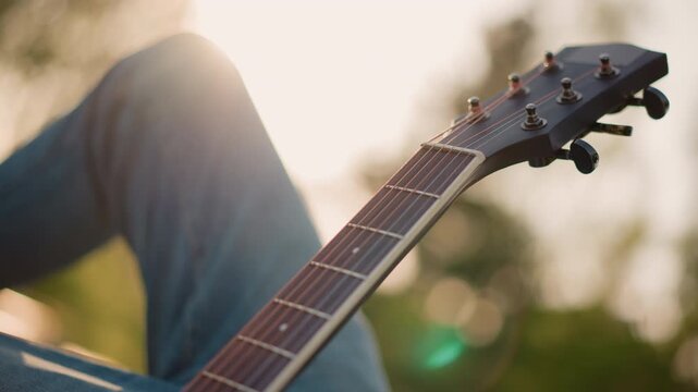 Guitarist relaxing outdoors. Casual musician seated in park. Serene scene of musician practicing with gentle sunlight. Peaceful image of artist playing guitar on park bench during sunset