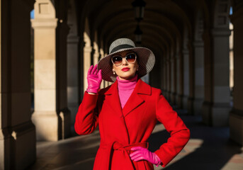 Naklejka premium Woman in red coat and pink gloves standing confidently in a beautiful arched hallway with columns and natural light