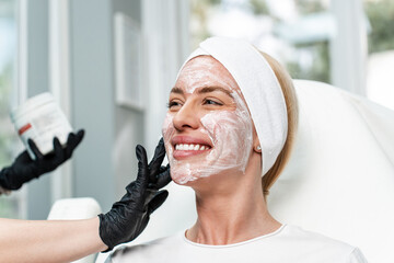 A professional aesthetician in black gloves applies a cooling numbing gel to a smiling woman's face in a bright clinic before a Botox or dermal filler procedure.