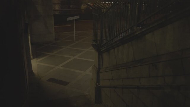 A couple descends an urban stairway and then starts running into a narrow underpass in a moody nighttime scene.