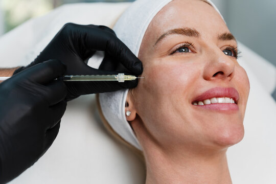 A smiling woman receives a professional facial filler injection in a modern aesthetic clinic. The procedure is performed by a doctor wearing black gloves.