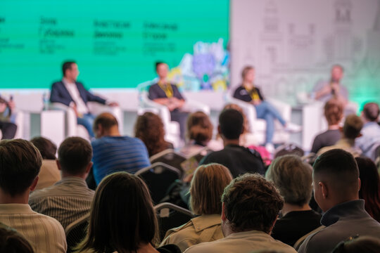 Panel discussion at conference with speakers on stage and attentive audience. Bright backdrop creates energetic mood.