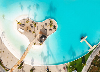 Aerial drone view artificial tropical lagoon in island beach surrounded by turquoise water, with palm trees and straw umbrellas. Vacation, summer holiday and relaxation