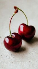 Close up of two ripe red cherries with long green stems resting on a light textured surface with soft natural light highlighting their glossy skin and fresh appearance