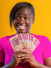 A woman in a vibrant pink shirt smiles joyfully while fanning out several 200 Ghanaian Cedi banknotes against a bright yellow background. She looks aside with a playful, excited expression.