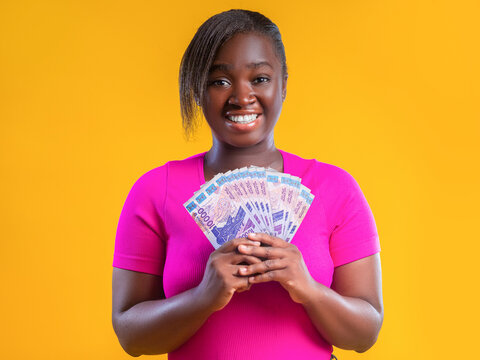 A joyful Black woman in a bright pink t-shirt holds a fan of West African CFA franc banknotes against yellow backdrop. The purple and brown hues of the notes signify high value 10000 CFA denomination