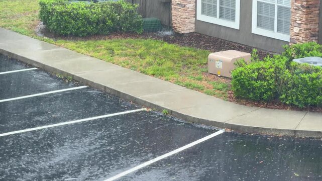 Heavy rain pours onto an empty parking lot and wet sidewalk beside a building. Water splashes across the dark asphalt while grass and bushes soak in the storm.