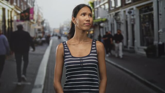 Thai woman with bare shoulder and visible collarbone wearing a striped tank top, looking to the side on a street lined with storefronts and pedestrians; quiet contemplation.