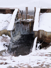 Small weir on artificial reservoir partially frozen in winter with flowing water beneath ice. Concept of seasonal infrastructure, contrast of ice and water, cold weather conditions.