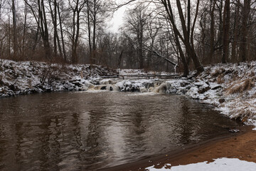 Wodospad na niewielkiej rzece. Jest zima. Brzegi pokryte są warstwą śniegu. Rzekę miejscami pokrywa lód. Wzburzona woda na wodospadzie przepływa pomiędzy lodowymi czapami. © boguslavus