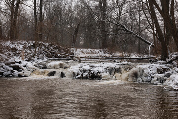 Wodospad na niewielkiej rzece. Jest zima. Brzegi pokryte są warstwą śniegu. Rzekę miejscami pokrywa lód. Wzburzona woda na wodospadzie przepływa pomiędzy lodowymi czapami. © boguslavus