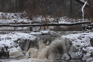 Wodospad na niewielkiej rzece. Jest zima. Brzegi pokryte są warstwą śniegu. Rzekę miejscami pokrywa lód. Wzburzona woda na wodospadzie przepływa pomiędzy lodowymi czapami. © boguslavus