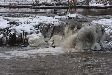 Wodospad na niewielkiej rzece. Jest zima. Brzegi pokryte są warstwą śniegu. Rzekę miejscami pokrywa lód. Wzburzona woda na wodospadzie przepływa pomiędzy lodowymi czapami. © boguslavus