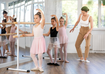 Adult woman teacher helping group of girls dancers practicing plie movement at barre in studio