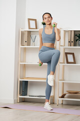 Naklejka premium Fitness woman performing a knee raise with dumbbells at home, showcasing strength training, focused routine, and athletic movement in a tidy living space.