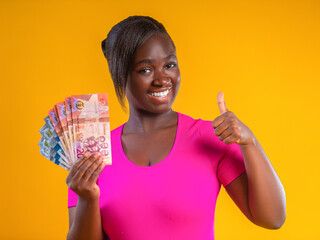 A young woman with a dark complexion smiles brightly, holding a fan of Ghanaian Cedi banknotes and giving a thumbs-up. She wears a vibrant pink shirt against a solid, bold yellow background.