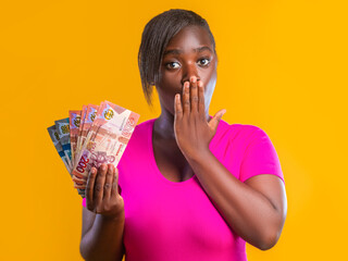 A woman in a vibrant pink shirt expresses shock, covering her mouth while holding a fan of Ghanaian Cedi banknotes against a bright yellow background. The image conveys surprise and financial success.
