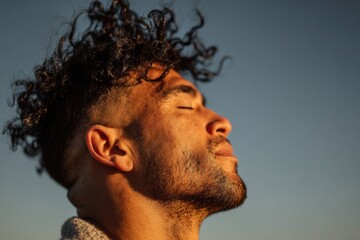 Fototapeta premium Man with eyes closed stands in warm sunlight as wind blows his curly hair across his face creating a calm moment