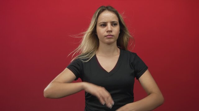 Young blonde woman standing with hands on hips wearing black v neck shirt and a slight squint in red studio; frustration.