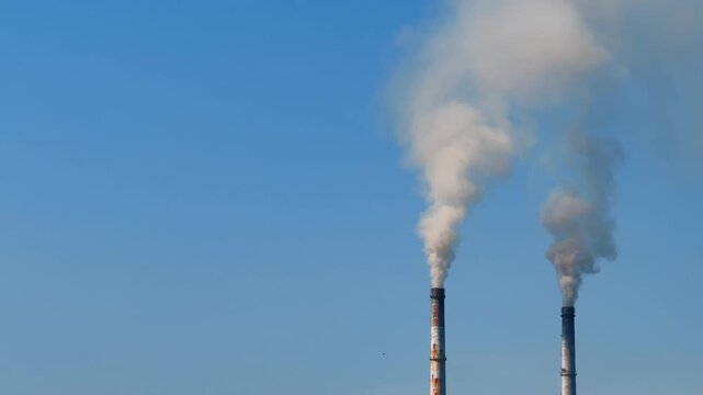 Smoke rises from factory chimneys in a clear blue sky during daylight hours. Factory smoke pours from two tall chimneys into the blue sky in a typical urban setting.