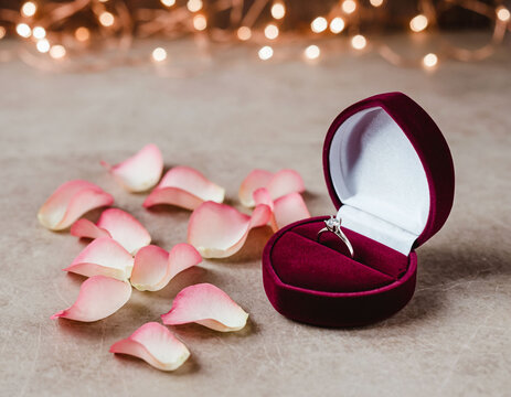 Anillo de compromiso en estuche rojo sobre p&eacute;talos de rosa con luces bokeh, propuesta rom&aacute;ntica
