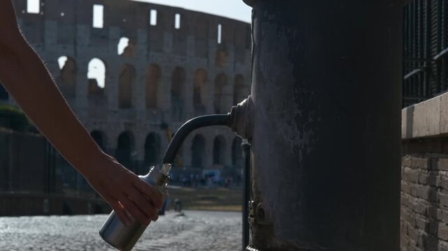 Tourist filling reusable bottle at public fountain near colosseum. Close-up of hands filling a metal flask with fresh water from a traditional roman drinking fountain, with the famous colosseum